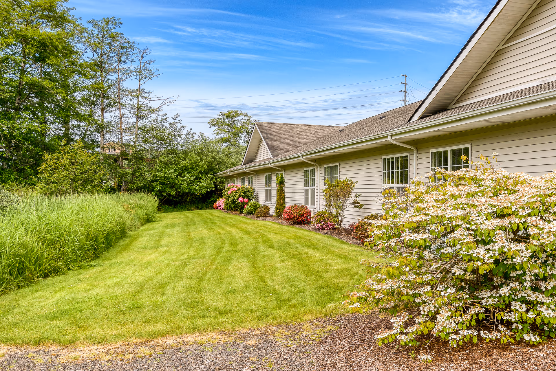 Well-maintained lawn and landscaped side of a single-story memory care building with shrubs and windows under a blue sky.