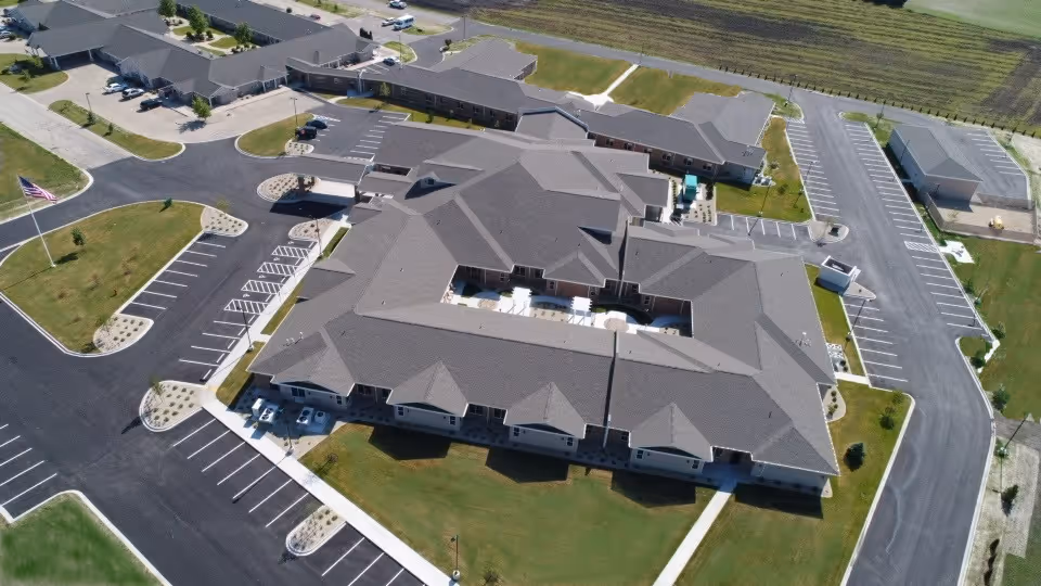 Aerial view of Liberty Village of Rochelle, showing a large senior living facility building with multiple wings, surrounded by parking lots, green lawns, and walkways. The building has a gray roof and is situated in a spacious area with some landscaping and an American flag visible near the parking lot.