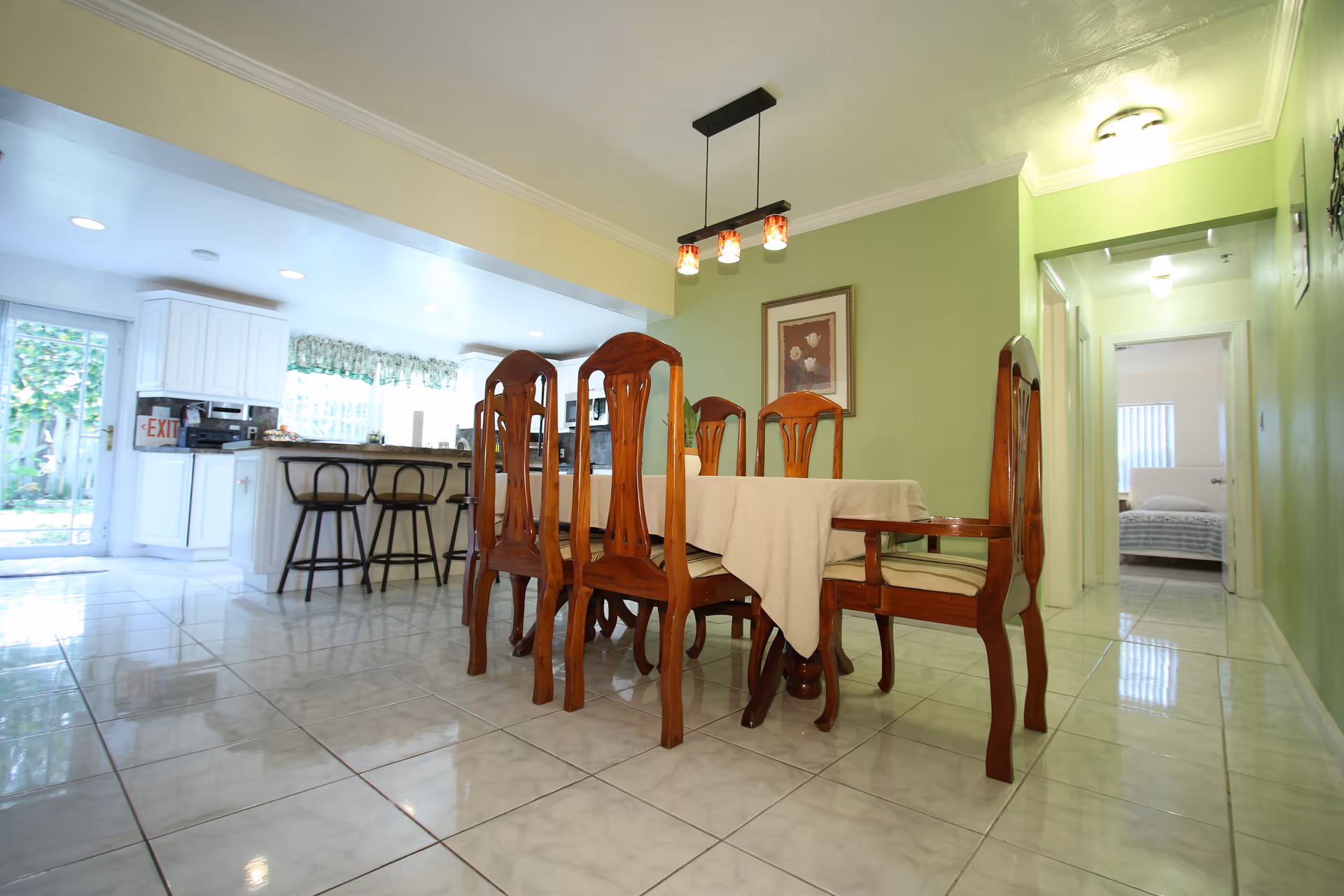 A bright dining area with a wooden dining table covered with a beige tablecloth and six matching wooden chairs. The room has light green walls and a tiled floor. A modern light fixture with three hanging lights is above the table. In the background, there is a kitchen with white cabinets, a counter with three black bar stools, and a glass door leading outside. A hallway to the right leads to a bedroom with a bed visible through the doorway.