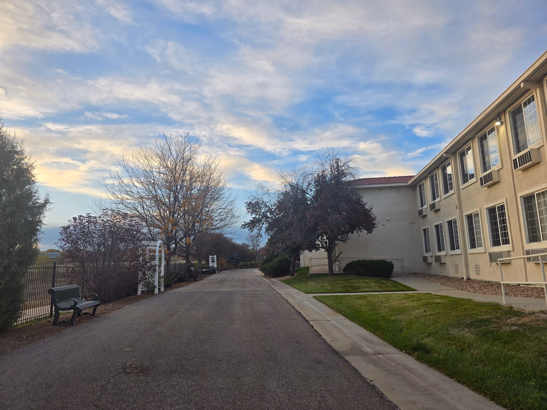 Driveway lined with benches and trees leading past the exterior of a multi-story retirement community building under a blue sky.