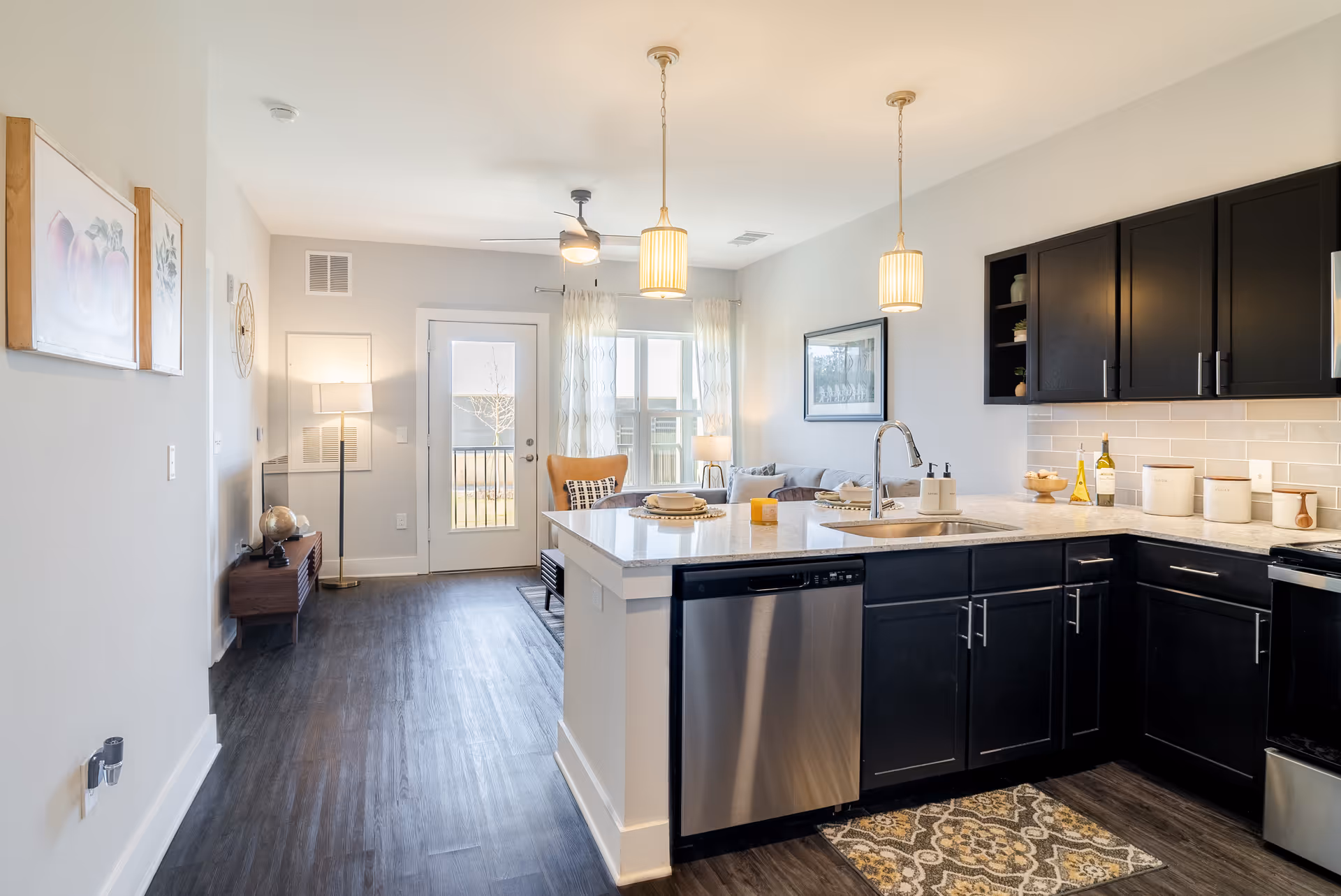 Bright open-plan kitchen with a central island, dark cabinets, stainless dishwasher, and a view into the adjacent living area and exterior door.
