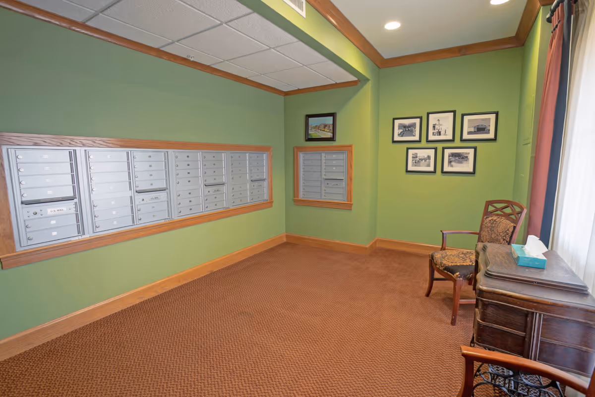A mailroom with green walls and brown carpet featuring multiple silver mailboxes mounted on the wall. There is a wooden chair with patterned upholstery and a wooden table with a tissue box near a window with curtains. Several framed black and white photographs hang on the wall.