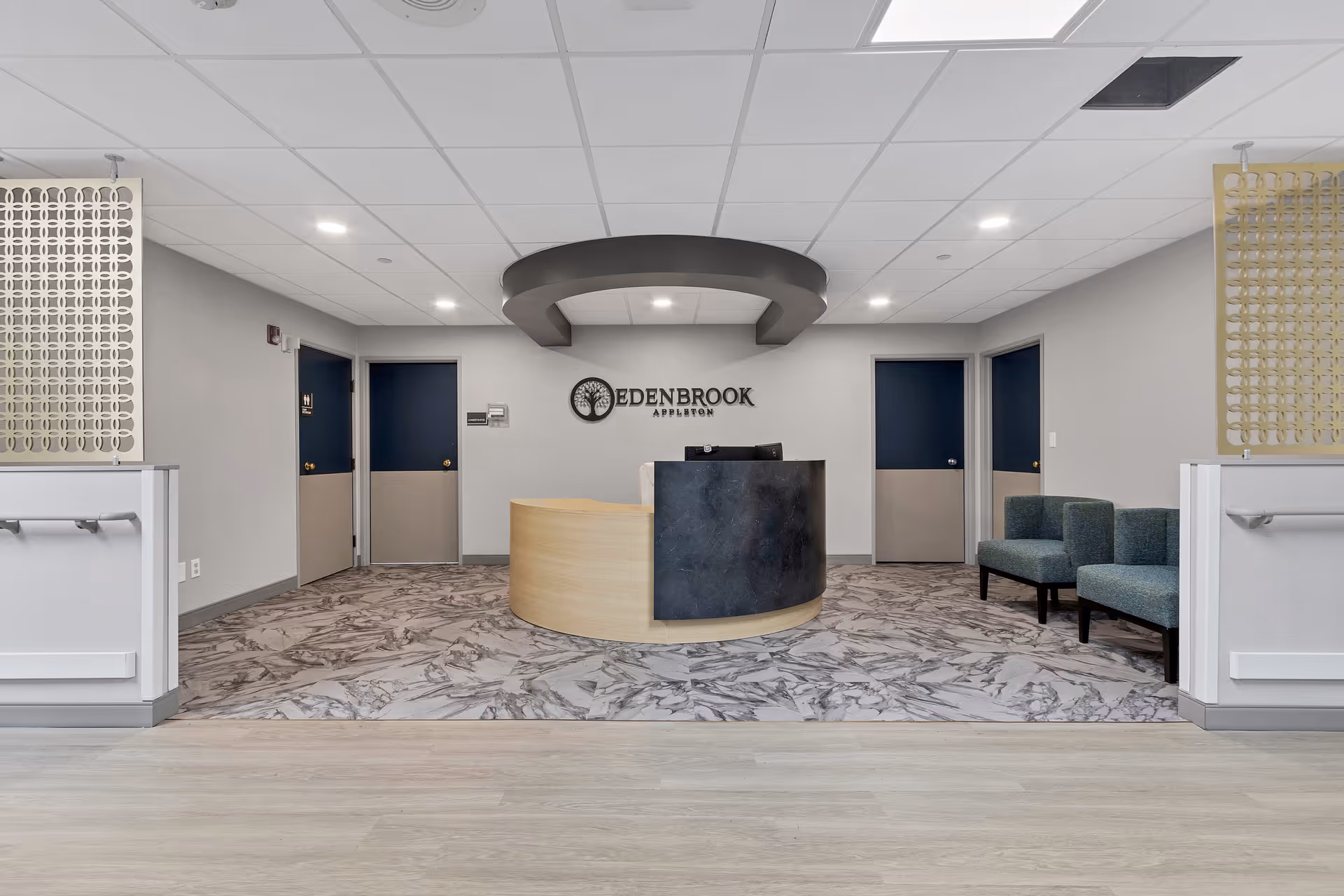 Reception area of Edenbrook Appleton with a curved wooden and black reception desk centered on a marble-patterned floor. Behind the desk is a wall with the Edenbrook Appleton logo. The room has a light gray ceiling with recessed lighting, light gray walls, and blue-gray doors. To the right, there are two green upholstered chairs. Decorative panels are on either side of the reception area.