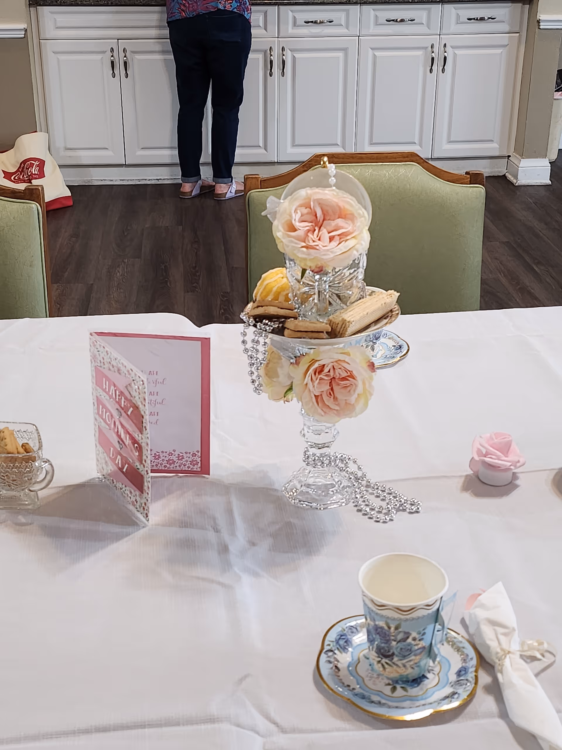 A table set with a white tablecloth featuring a decorative centerpiece with pink roses, a glass dish holding cookies, a floral teacup and saucer, a napkin with a ring, and a greeting card that says 'Happy Mother's Day'. In the background, a person stands facing white kitchen cabinets.
