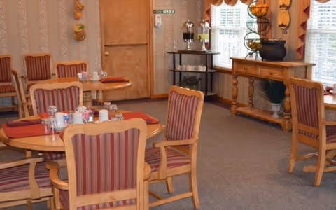 A dining room with round wooden tables and striped cushioned chairs. Tables are set with red placemats, salt and pepper shakers, and glassware. There is a wooden sideboard with decorative items and a beverage station with coffee dispensers near a door labeled 'Food Service'. Windows with blinds and valances allow natural light into the room.