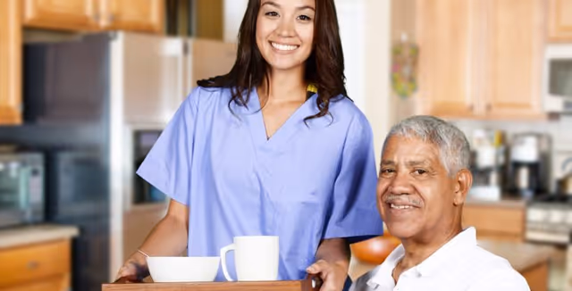 A smiling caregiver in scrubs stands behind an older man seated at a kitchen table with a tray holding a mug and bowl.