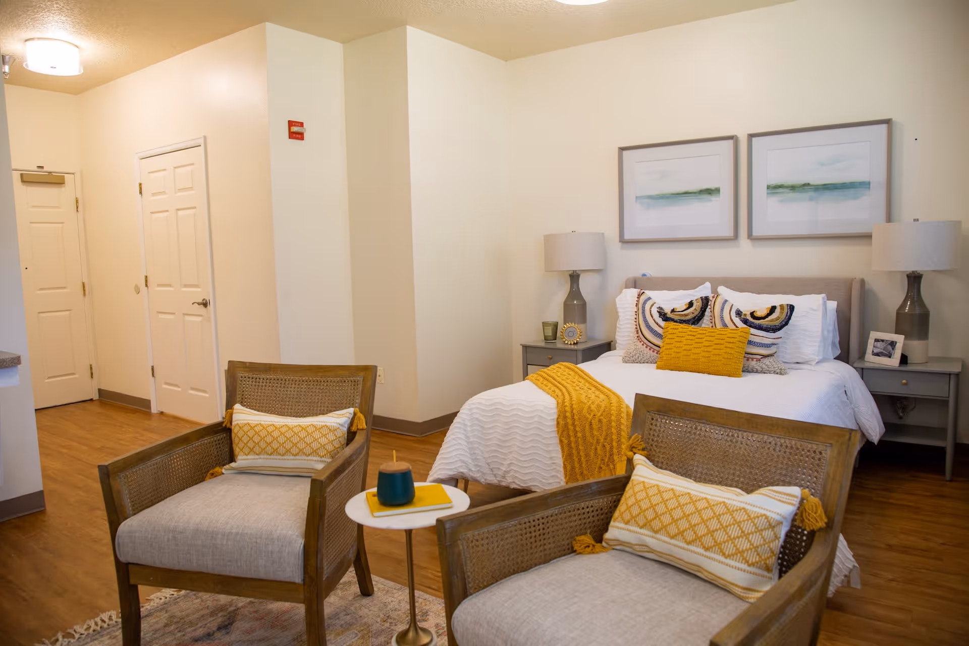 A cozy bedroom with a neatly made bed featuring white bedding and decorative pillows in yellow and patterned designs. Two matching bedside tables with lamps flank the bed. In front of the bed are two cushioned armchairs with yellow and white patterned pillows, separated by a small round side table holding a candle and a book. The room has wooden flooring, light-colored walls, and two framed landscape paintings above the bed. A hallway with closed doors is visible in the background.