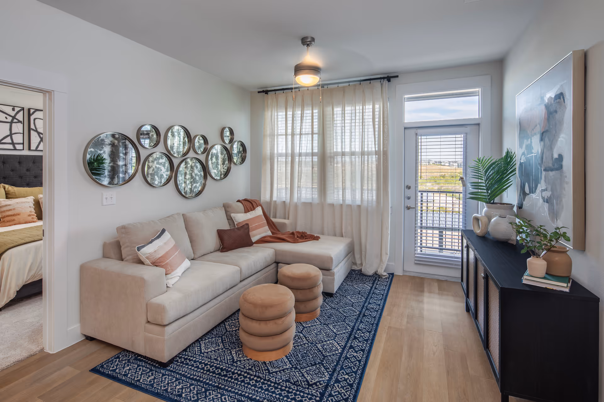 A cozy living room with a beige sectional sofa adorned with striped and solid pillows, two round beige ottomans on a blue patterned rug, a black console table with decorative vases and plants, a large abstract painting on the wall, and a window with sheer curtains next to a glass door leading outside. A bedroom with a bed and pillows is partially visible through an open doorway.