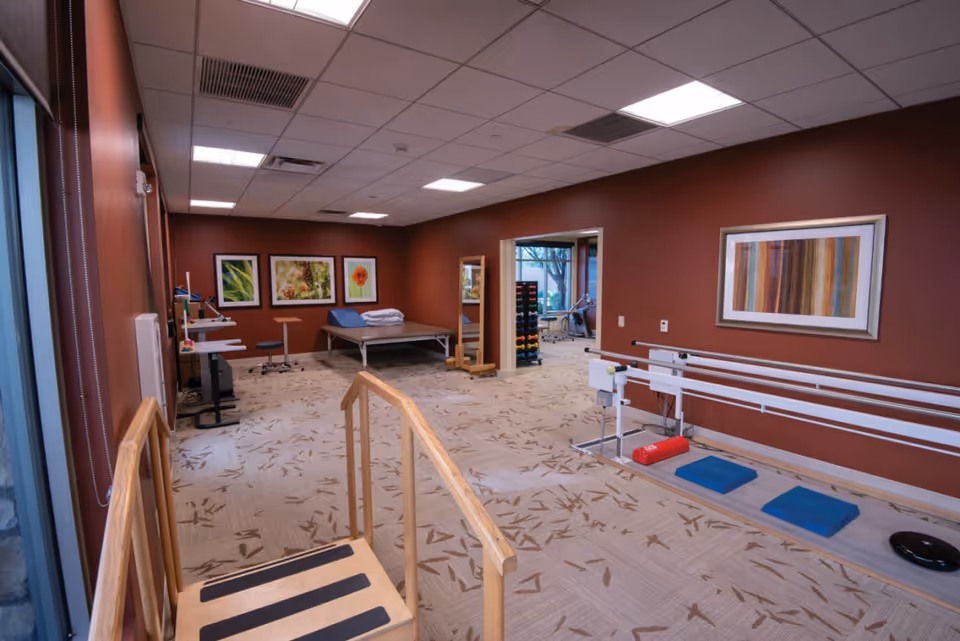 A rehabilitation or physical therapy room with wooden parallel bars, exercise mats, a treatment table with pillows, exercise equipment, and framed artwork on the walls. The room has a patterned carpet and a ceiling with recessed lighting.