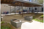 Outdoor patio area at Riverside Rest Home with a covered seating area featuring multiple tables and chairs. There are two rectangular planter boxes in the foreground and a small grassy area to the right.