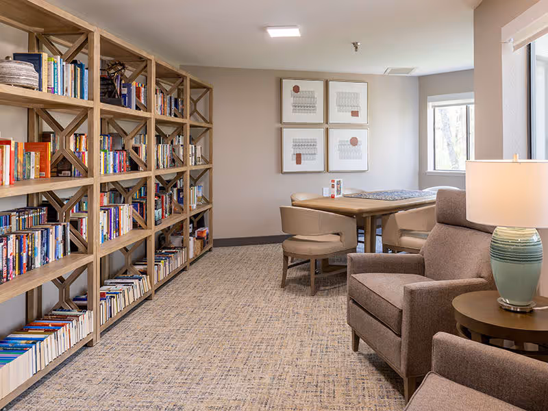 A cozy interior room with a large wooden bookshelf filled with books on the left side, a table with four beige chairs in the background, and two comfortable armchairs with a side table and a lamp on the right side. The room has carpeted flooring, neutral-colored walls, and framed artwork on the wall near a window.