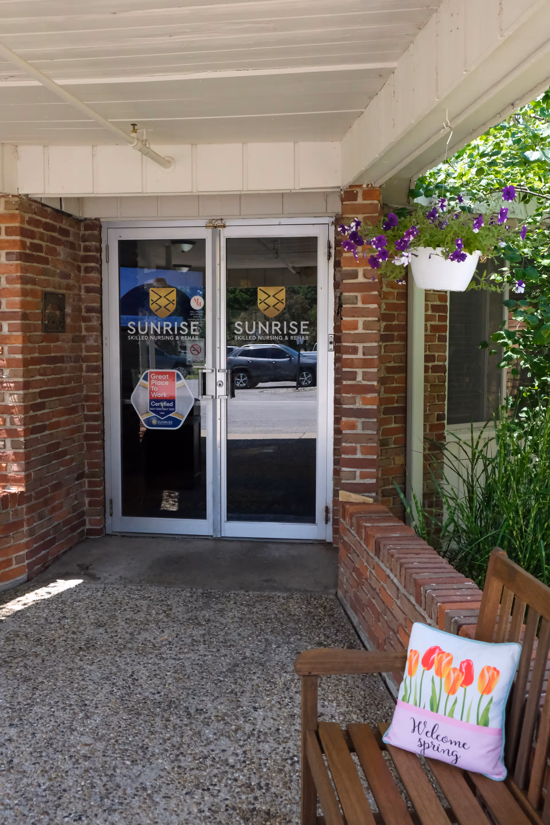 Entrance to Sunrise Skilled Nursing and Rehab facility with glass double doors featuring the facility's logo. There is a brick wall on either side of the doors, a hanging pot with purple flowers on the right, and a wooden bench with a colorful pillow that says 'Welcome spring'.