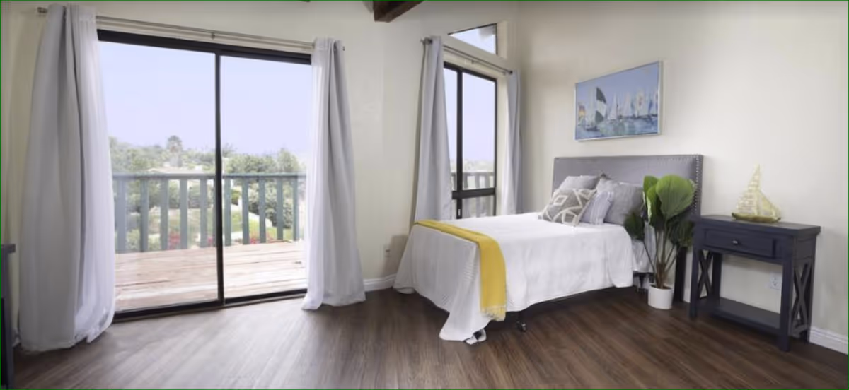 A bright bedroom with a bed covered in white bedding and a yellow throw blanket. The bed has several decorative pillows and a gray upholstered headboard. Next to the bed is a dark wooden nightstand with a decorative sailboat and a potted plant. The room has large sliding glass doors with white curtains that open to a balcony with a view of greenery outside. A painting of sailboats hangs on the wall above the bed.