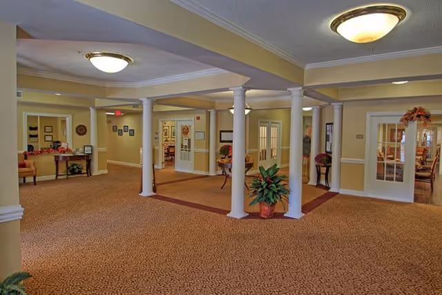 Interior view of a senior living facility common area with beige walls, carpeted floors, white columns, and ceiling lights. There are several doorways leading to other rooms, some with glass-paneled doors. The space is decorated with plants and small tables with flowers.