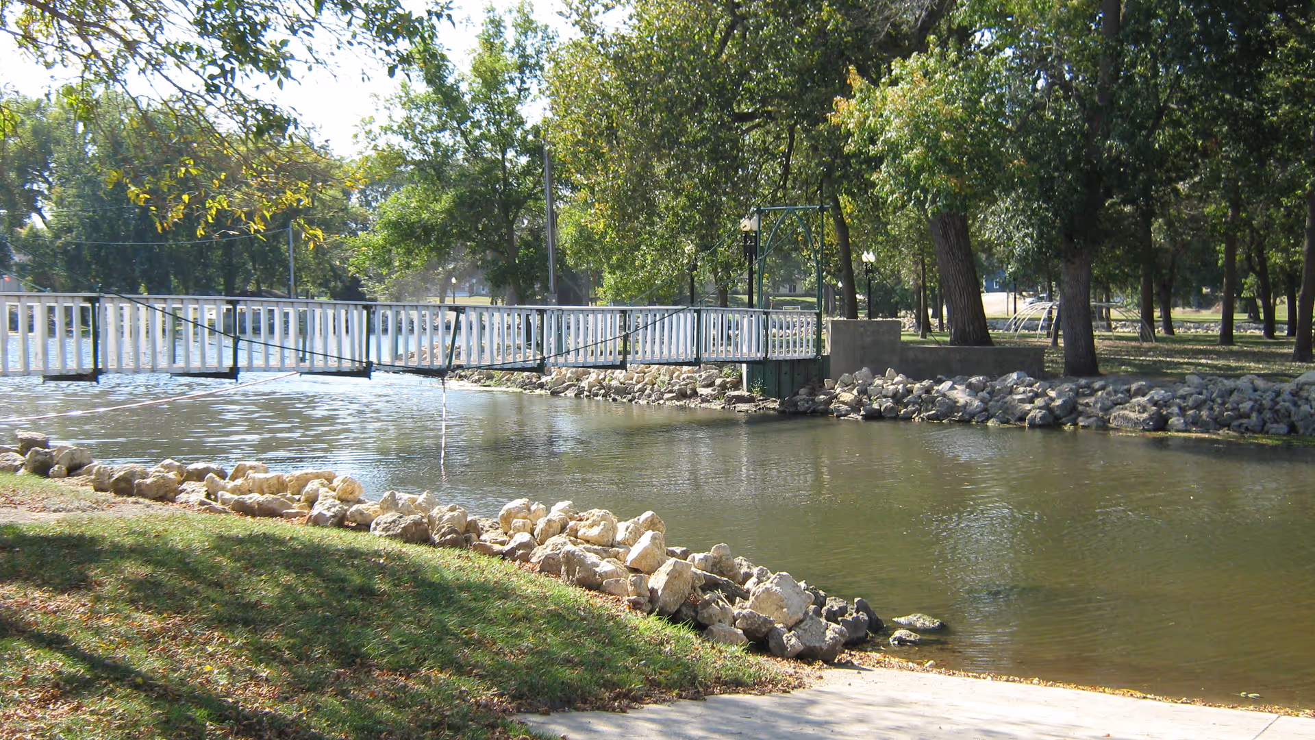 A peaceful outdoor scene featuring a small river or pond with a white pedestrian bridge crossing over it. The area is surrounded by green grass, rocks lining the water's edge, and numerous trees providing shade in the background.