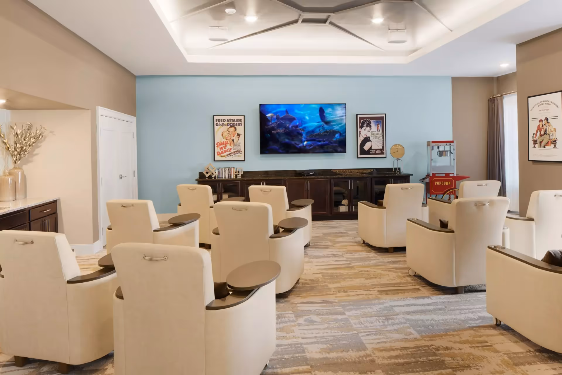 A cozy media room with beige armchairs arranged in rows facing a large flat-screen TV mounted on a light blue wall. The room has a popcorn machine in the corner, framed vintage movie posters on the walls, and a dark wooden cabinet beneath the TV. The carpet has a patterned design, and the ceiling features recessed lighting.