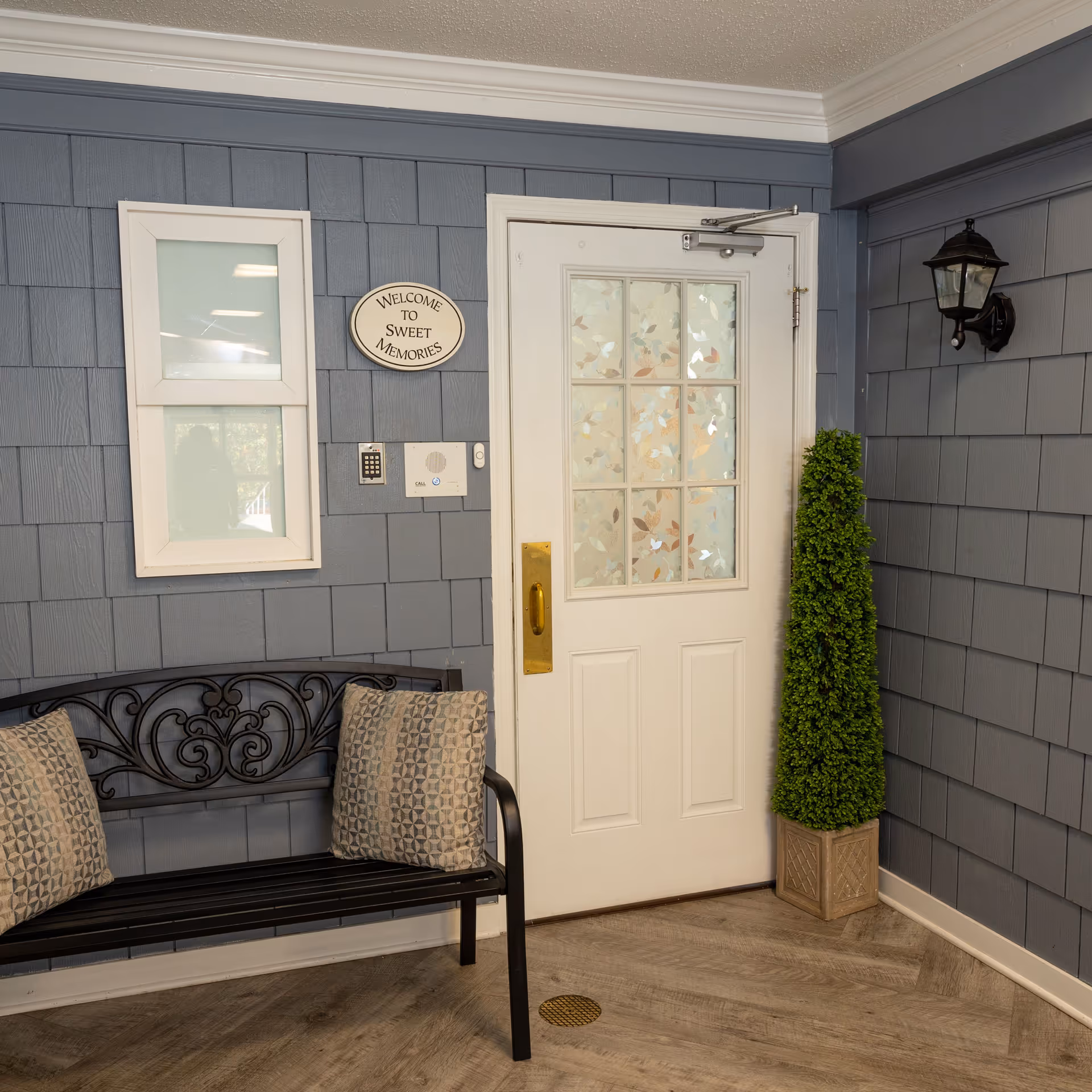 Interior entryway showing a white paneled door with frosted window, a decorative black bench with pillows, and a potted topiary against blue shingle-style walls.
