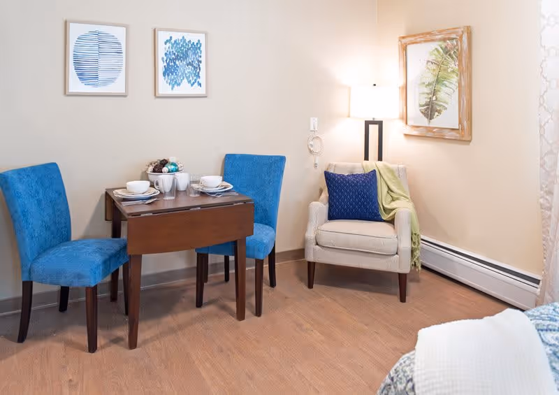 A cozy interior corner featuring a small wooden dining table set for two with blue upholstered chairs, a beige armchair with a blue pillow and green throw, a floor lamp, and framed artwork on the walls.