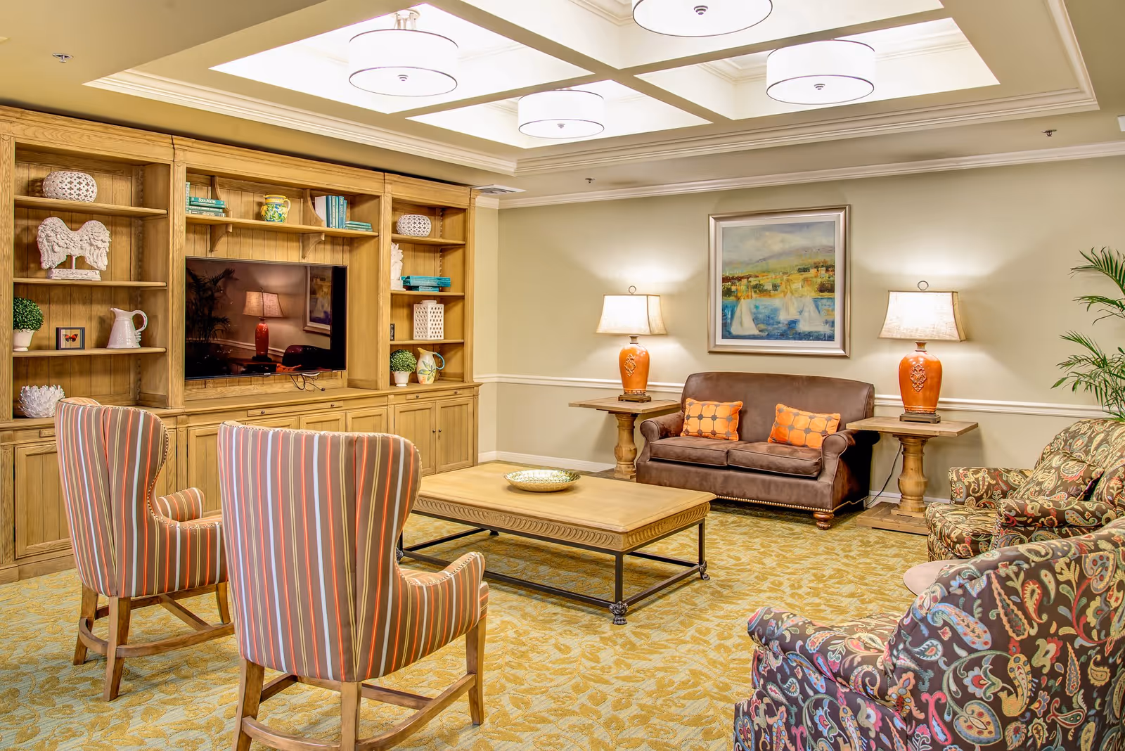A cozy living room with a wooden entertainment center holding a flat-screen TV and decorative items. The room features a brown leather loveseat with two orange patterned pillows, two striped armchairs, and two floral patterned armchairs. There are two wooden side tables with matching orange lamps, a large wooden coffee table, a framed painting on the wall, and a potted plant in the corner. The ceiling has recessed lighting with three round fixtures.