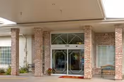 Entrance of a building with a covered drop-off area supported by brick columns. Double glass doors are centered under the covered area with windows on either side. There is a bench to the right and some potted plants near the entrance.