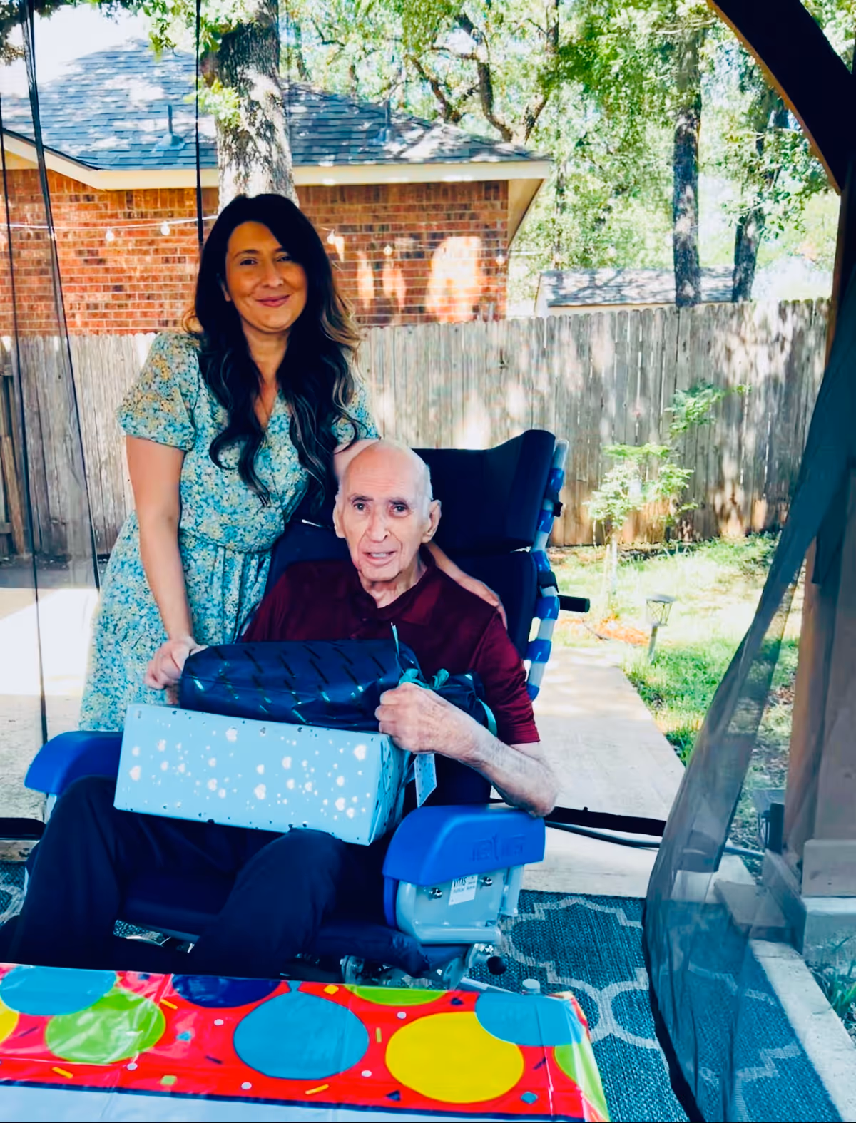 An elderly man sitting in a blue wheelchair holding two wrapped gift boxes, with a smiling woman standing behind him outdoors in a backyard setting with a wooden fence, trees, and a brick house in the background.