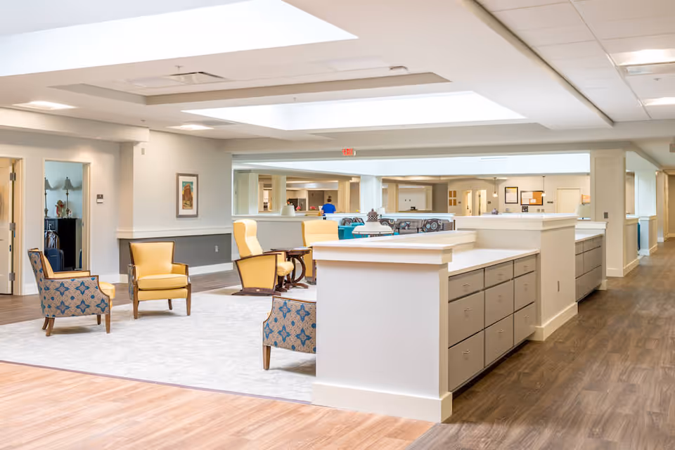 A bright and spacious common area in a senior living facility with comfortable seating including yellow and patterned armchairs arranged on a light-colored rug. The room features wood flooring, white walls, and a large skylight ceiling. There is a long counter with multiple drawers and cabinets, and an open hallway leading to other rooms.