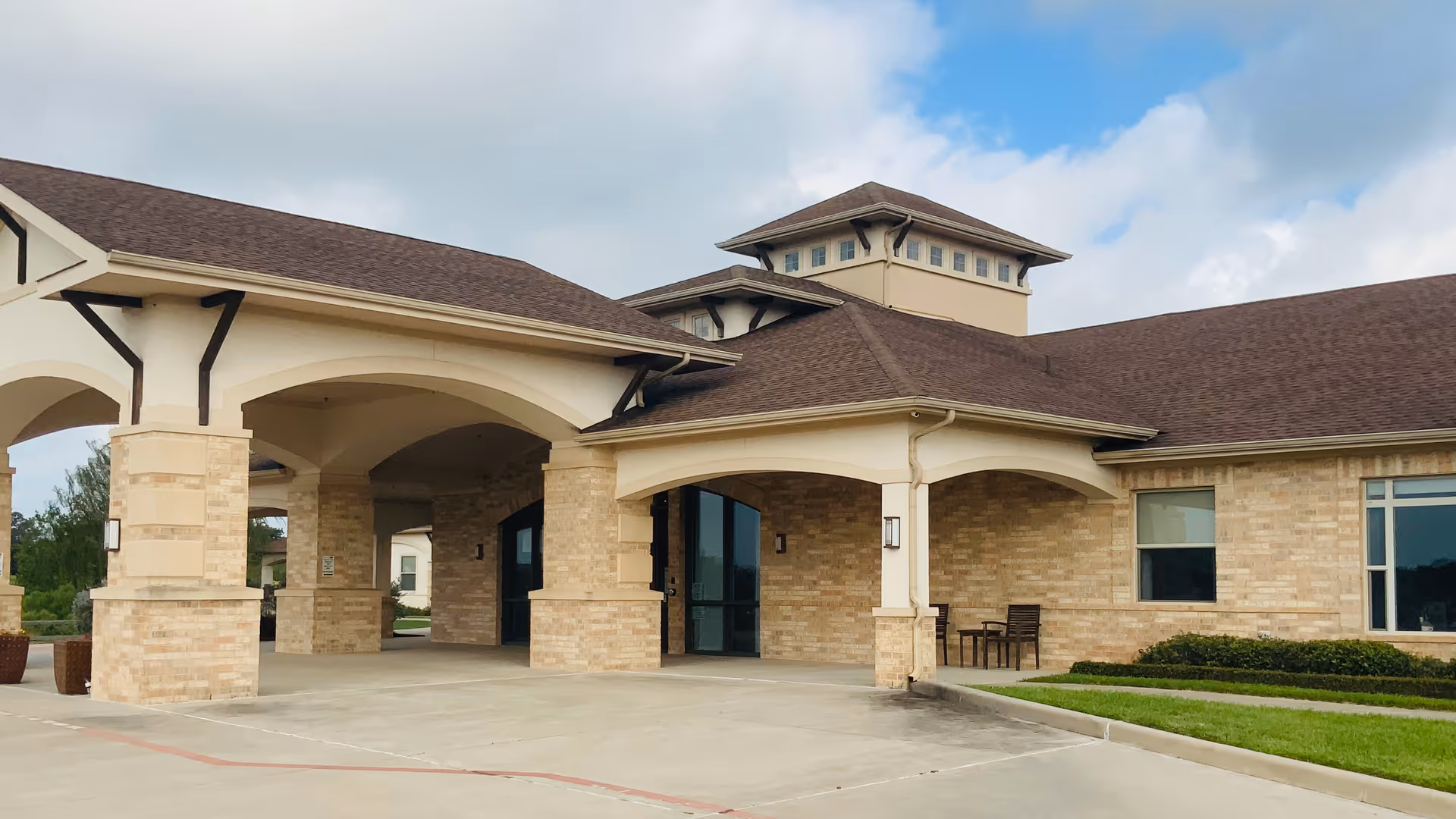 Front entrance of a brick senior living building with a covered porte-cochere and paved driveway under a partly cloudy sky.