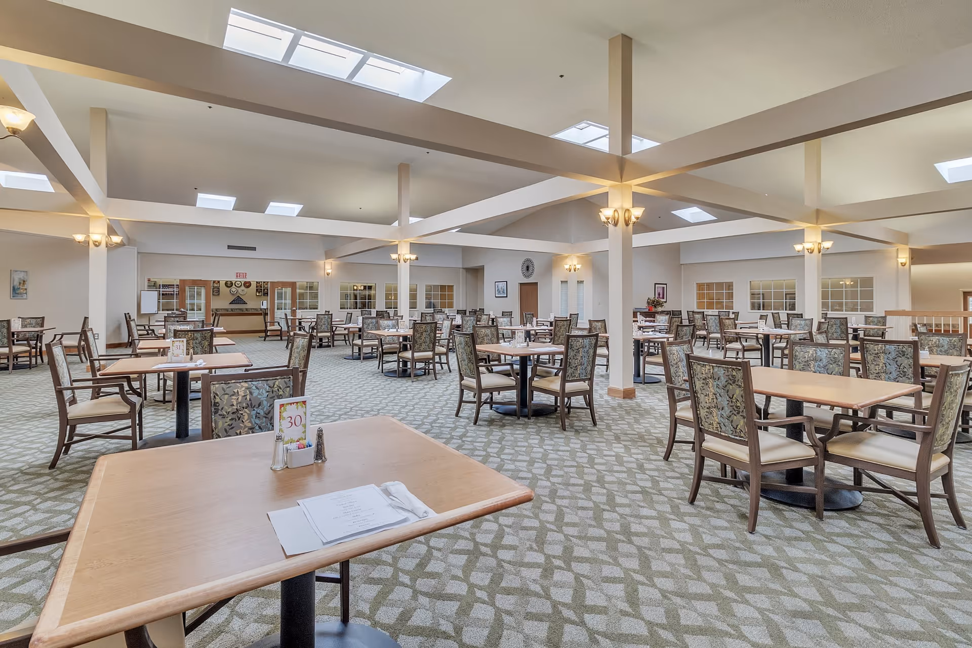 Spacious dining room with multiple wooden tables and cushioned chairs arranged neatly. The room has a patterned carpet, beige walls, and several skylights allowing natural light to brighten the space. Wall sconces provide additional lighting, and there are decorative elements on the walls.