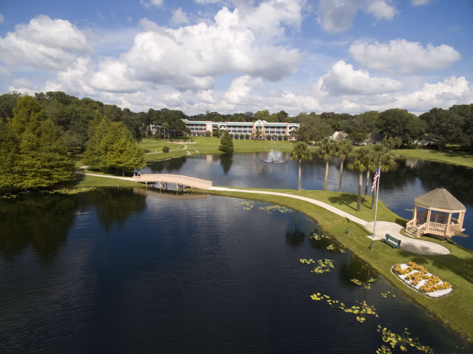 A scenic view of John Knox Village of Central Florida featuring a large pond with a fountain, a wooden bridge, palm trees, a gazebo, and a building in the background surrounded by greenery under a partly cloudy sky.