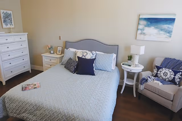 Bright bedroom with a light-blue quilted bed, decorative pillows, nightstands, an upholstered chair, dresser, and coastal wall art.