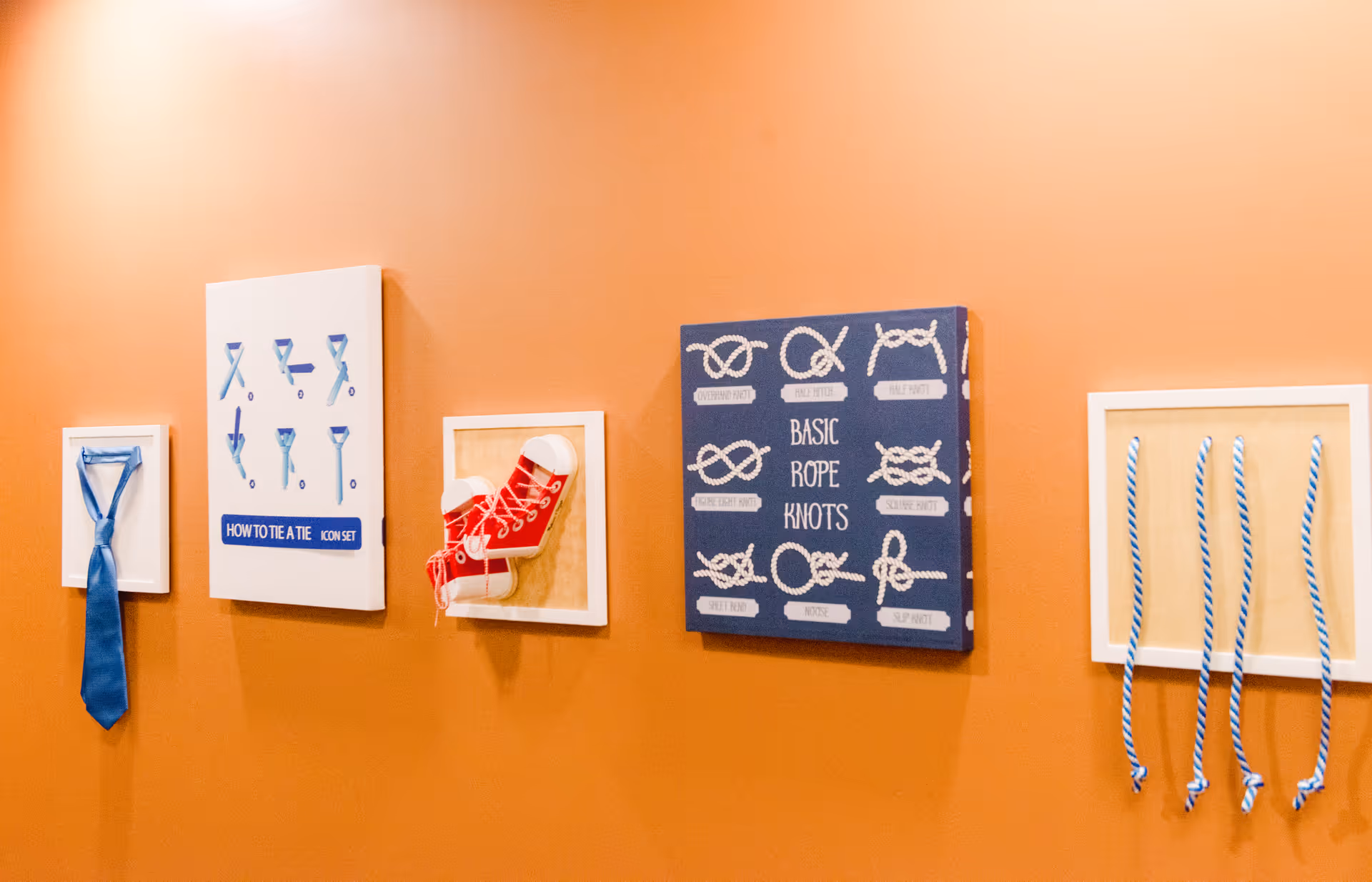 An orange wall with framed instructional displays showing how to tie a necktie, a pair of red sneakers, and various basic rope knots with labels. The displays include diagrams and actual objects mounted on the wall.