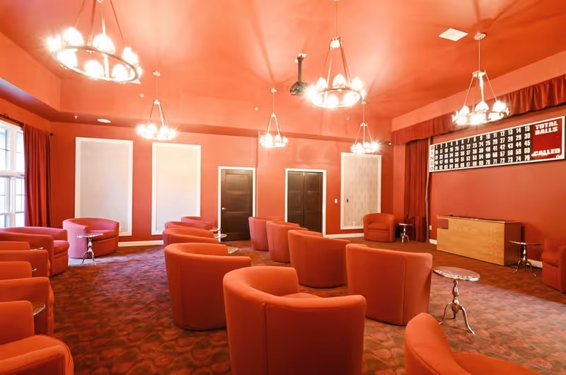 A spacious room with red walls, ceiling, and carpet, furnished with multiple red upholstered armchairs arranged in rows facing a bingo board on the wall. The room is lit by several chandelier-style ceiling lights, and there are small round side tables between some chairs. Two closed dark wooden doors are visible on the far wall.