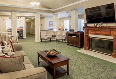 A cozy living room area in a senior living facility featuring a beige sofa with patterned cushions, a wooden coffee table with a decorative bowl, two upholstered armchairs with a small table between them, a wooden fireplace with a mounted flat-screen TV above it, and green patterned carpet flooring. The room is softly lit with ceiling lights and has a warm, inviting atmosphere.