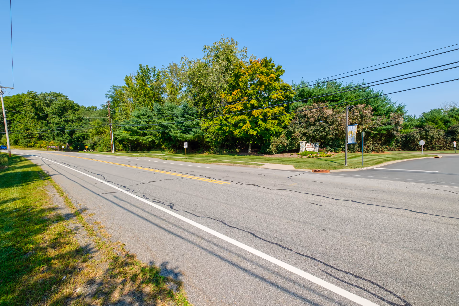 A wide two-lane road bordered by grass and trees with a landscaped entrance and sign to a community on the right.