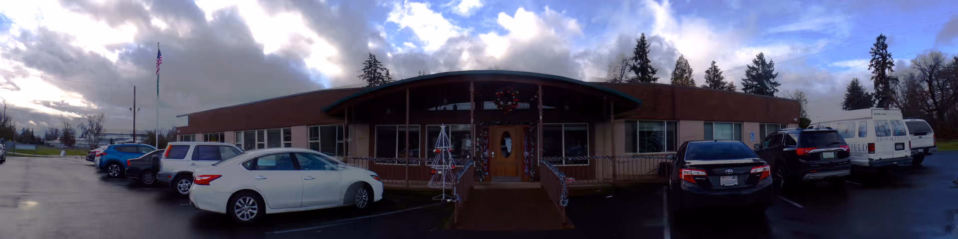 Exterior front view of a single-story brick building with a curved roof entrance decorated with holiday wreaths and lights. Several cars are parked in front of the building under a cloudy sky with patches of blue. An American flag is visible on a flagpole to the left.