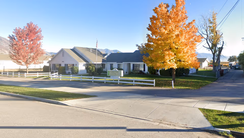 Exterior view of Country View Assisted Living facility with a white fence, green lawn, and trees with autumn foliage. The building is a single-story structure with a gray roof and an American flag on a flagpole in front. Mountains are visible in the background under a clear blue sky.