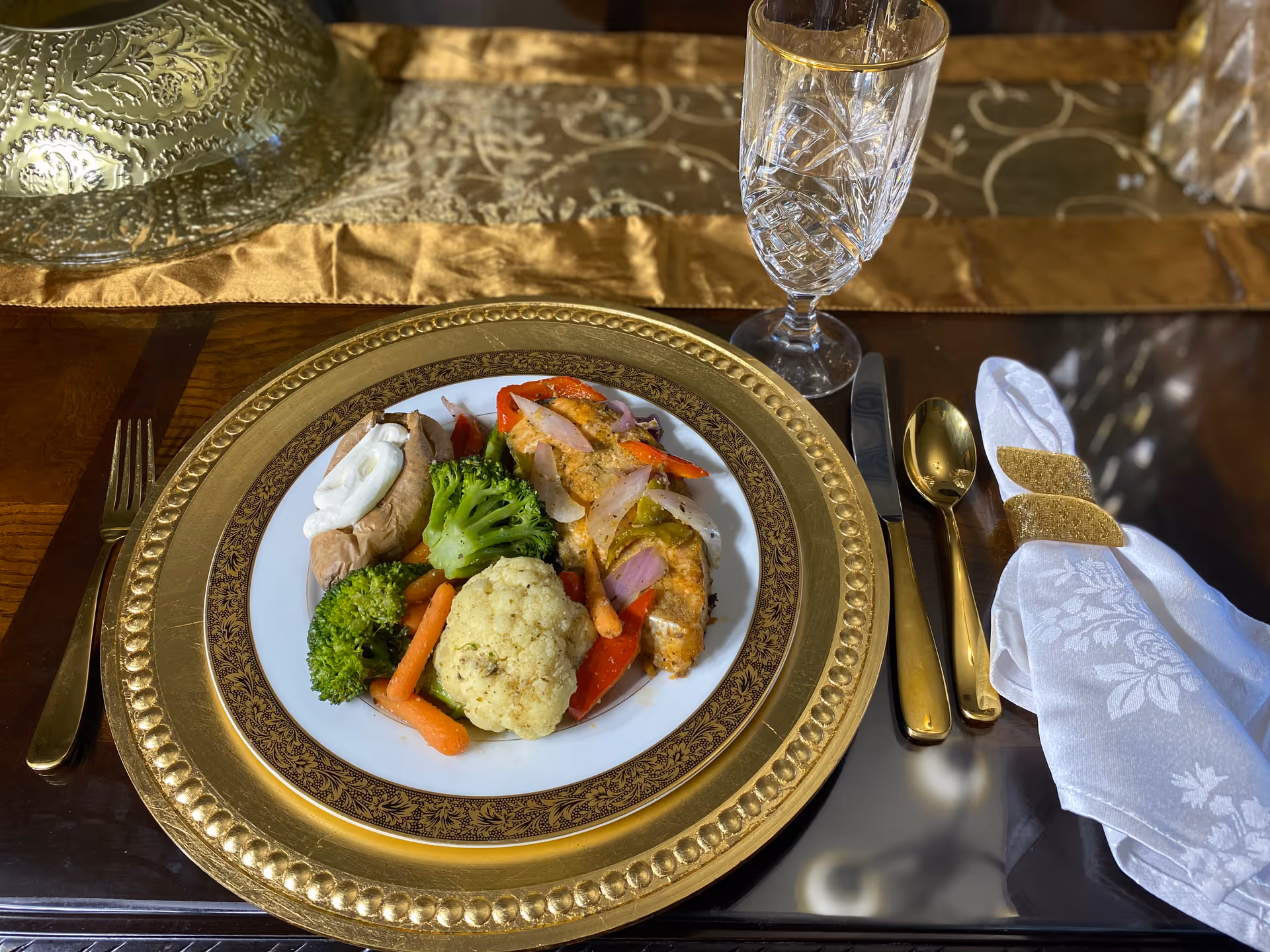 A plated meal on a decorative gold and white plate set on a gold charger plate. The meal includes cooked broccoli, cauliflower, baby carrots, red bell peppers, onions, a baked potato with sour cream, and a piece of seasoned chicken. The place setting includes a gold fork, knife, and spoon, a crystal water glass, and a white cloth napkin with a gold napkin ring, all arranged on a dark wooden table with a gold and patterned table runner.