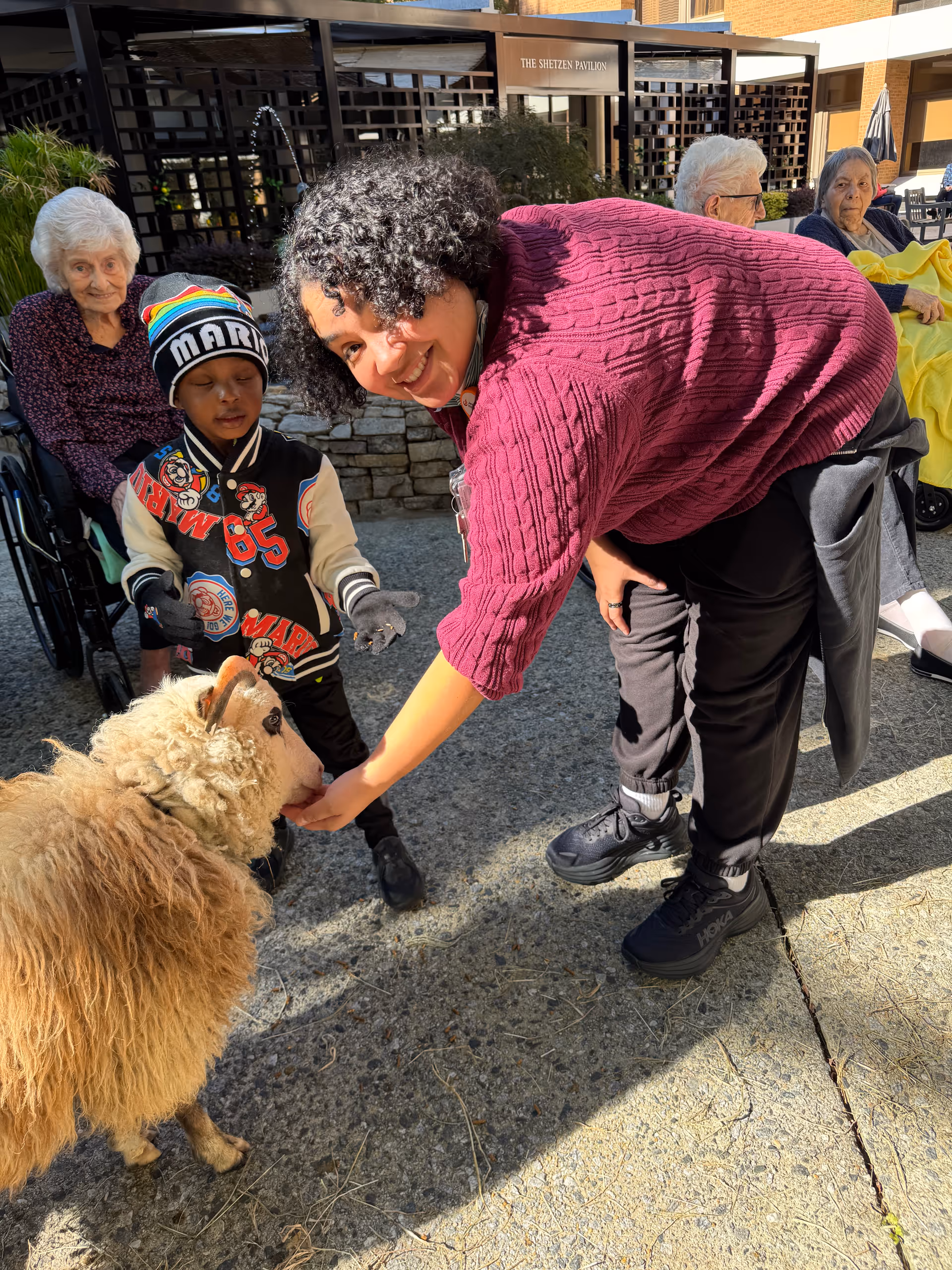 A woman in a purple sweater is bending down to feed a sheep while a young boy wearing a colorful Mario hat and jacket stands nearby. In the background, three elderly women are seated, two in wheelchairs and one on a bench, outside a building with a sign that reads 'The Shetzen Pavilion'.