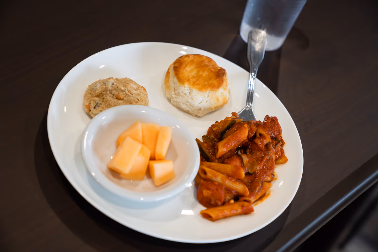 A white plate holding penne pasta in tomato sauce, a biscuit, a small muffin, and a bowl of cantaloupe with a glass of water on a dark table.