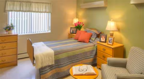 Cozy bedroom in Hillcrest Nursing Center featuring bed, nightstands, dresser, and chair.