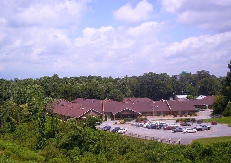 Aerial view of Westwood Hills Nursing and Rehabilitation Center surrounded by trees and greenery under a partly cloudy sky. The building has a brown roof and a parking lot with several cars parked.