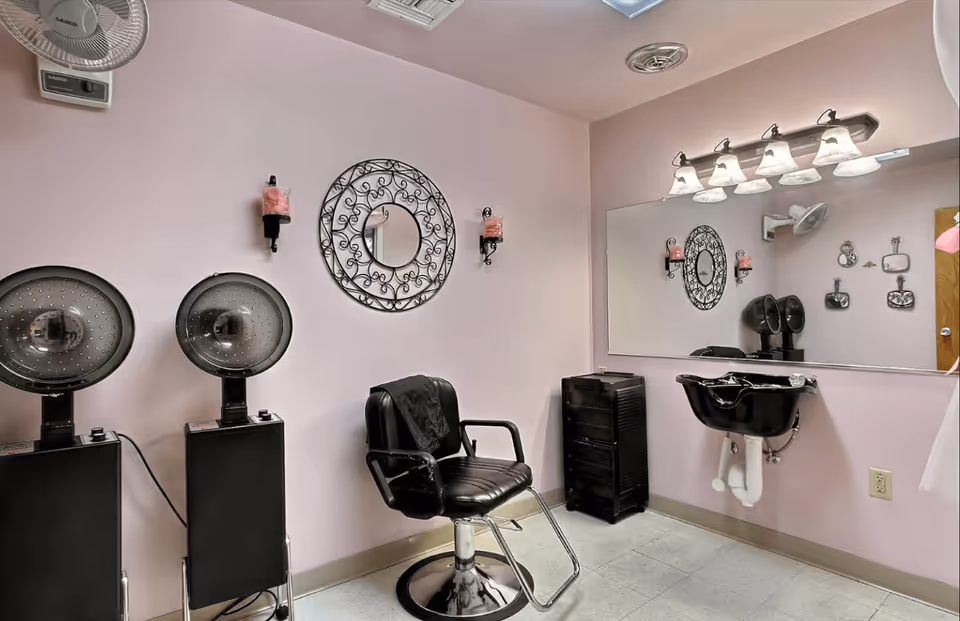 Interior of a small hair salon or beauty station with two black hair dryers, a black salon chair, a black sink for washing hair, a large wall mirror, and decorative wall accents including a round ornate mirror and candle holders on a light pink wall.