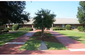 Exterior view of a single-story building with a beige roof and brick walls, surrounded by green grass, bushes, and trees. A paved walkway leads to the entrance, with a lamp post in the center of the path.