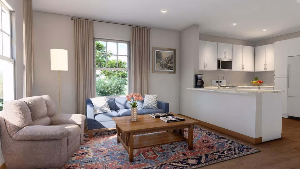 A bright and cozy living room area with a beige armchair, a blue sofa with two patterned pillows, a wooden coffee table with a vase of pink tulips and books, a floor lamp, and a large window with beige curtains. The room opens into a modern kitchen with white cabinets, a marble countertop island, a coffee maker, and a bowl of fruit.