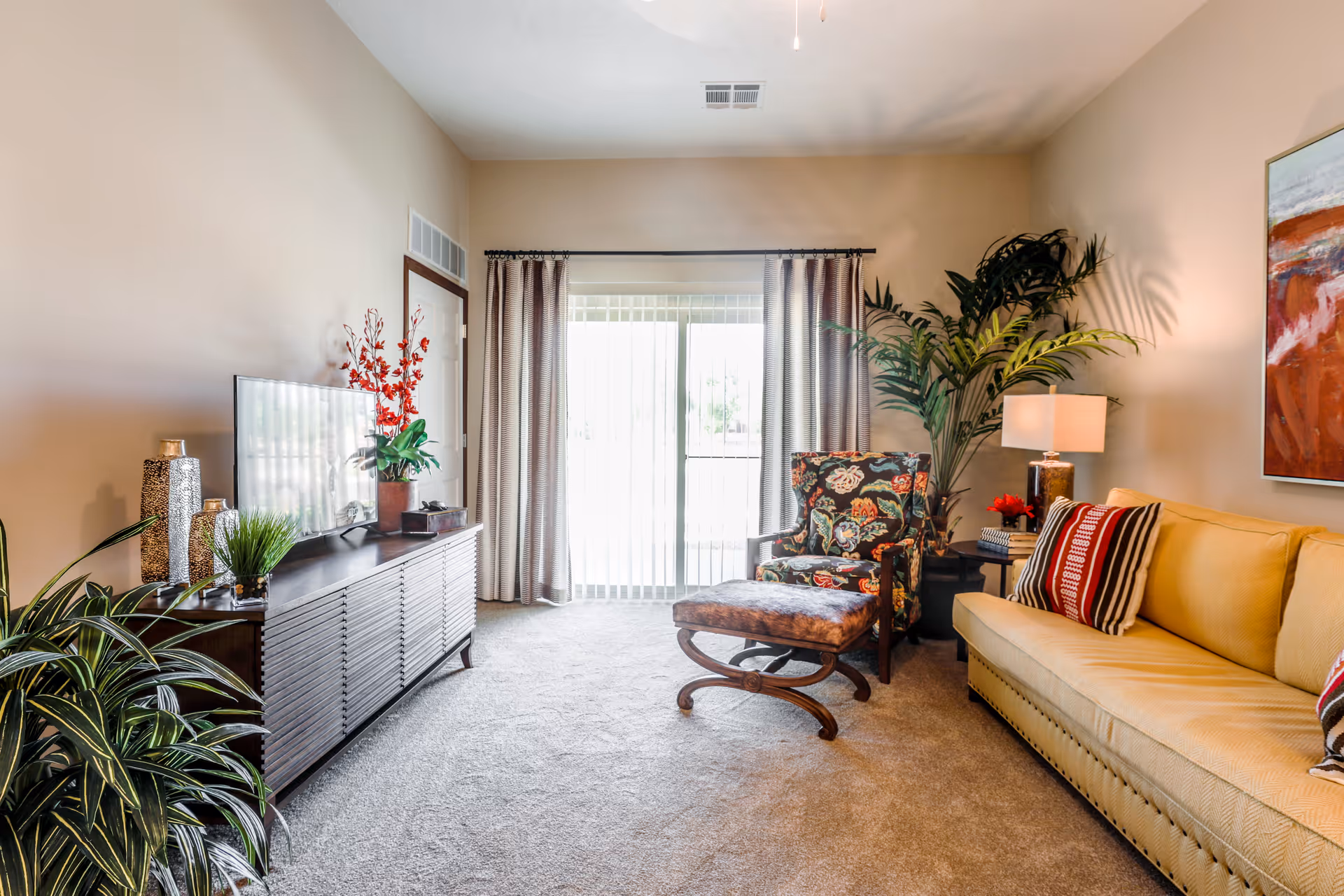 A bright living room with a beige sofa adorned with striped and patterned cushions, a floral upholstered armchair with matching ottoman, a wooden TV stand with a flat-screen TV and decorative plants, a side table with a lamp and a large potted plant near sliding glass doors covered with curtains.