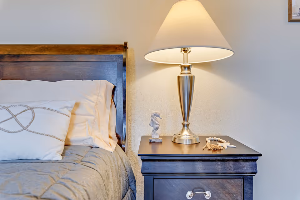 Bedside table with a lit lamp, small seahorse decoration and beads next to a wooden headboard and pillows.