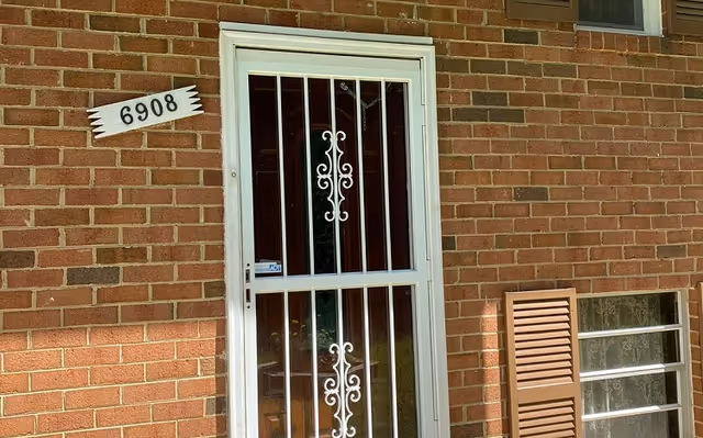 A brick exterior wall of a building with a white metal security door featuring decorative scrollwork. To the left of the door is a white house number sign displaying 6908. To the right of the door is a window with brown shutters, one shutter is partially open.