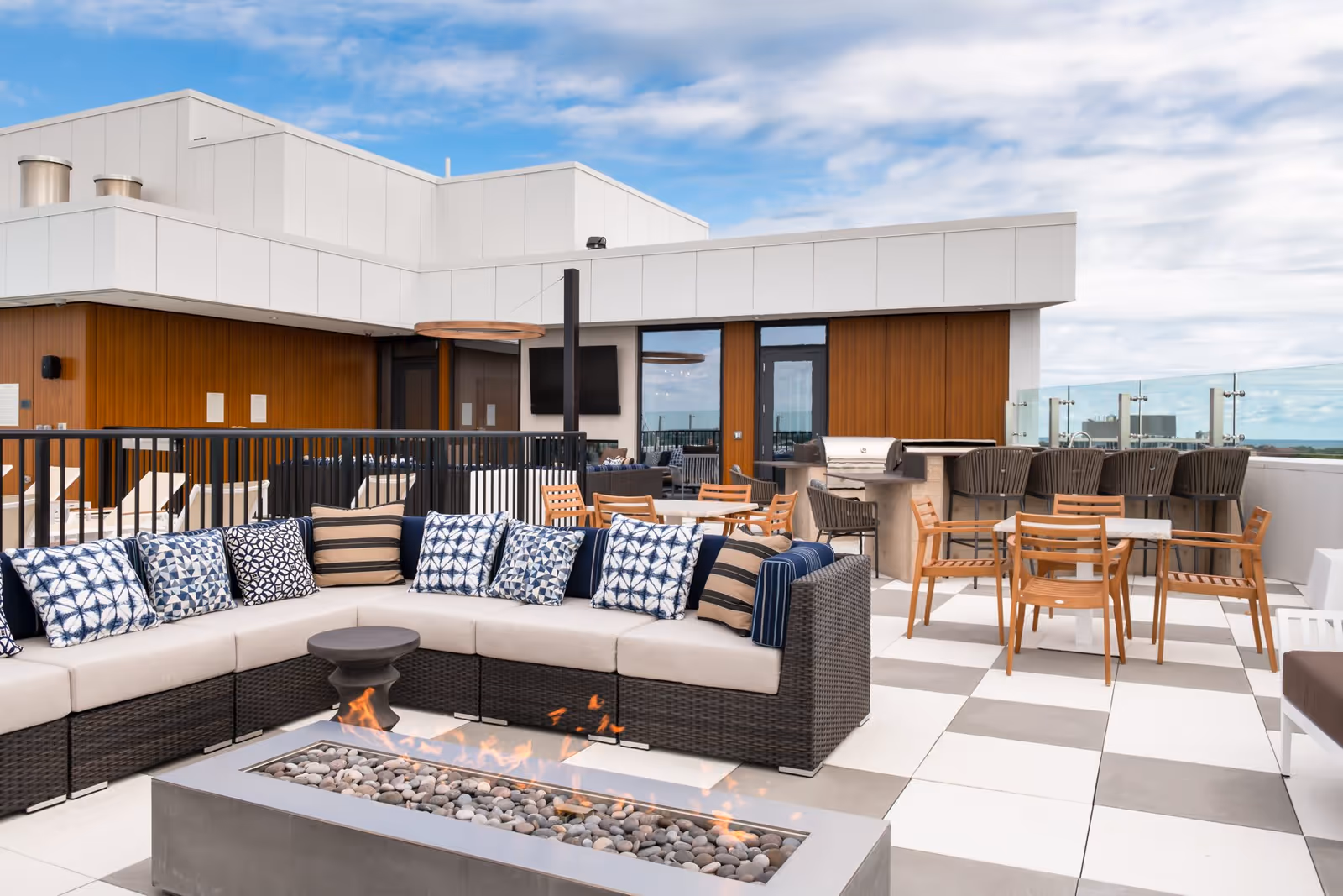 Outdoor rooftop patio area with a modern sectional sofa adorned with patterned cushions, a rectangular fire pit with flames, several wooden chairs around tables, a barbecue grill, and a bar counter with stools. The area is surrounded by glass railings and features a building with wood paneling and white exterior walls under a partly cloudy sky.