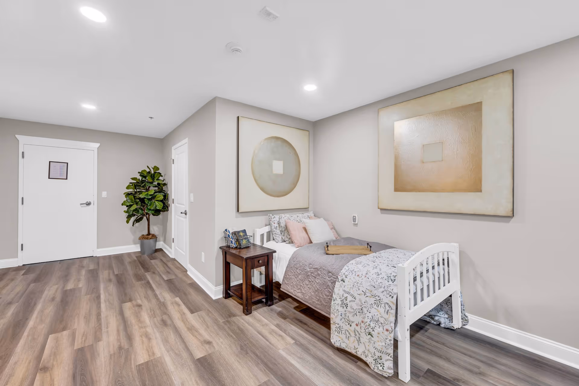A neatly arranged bedroom in a senior living facility featuring a single white bed with floral and solid color bedding, a wooden side table with books, two large abstract paintings on the walls, a potted plant in the corner, and light wood flooring.