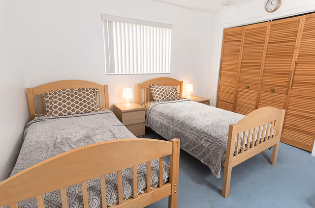 A simple bedroom with two wooden twin beds, a nightstand with lamps between them, a window with vertical blinds and a louvered closet.